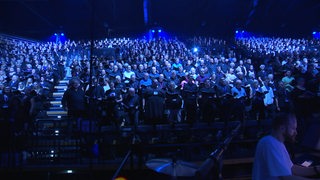 Personen singen im Chor. Sie sitzen in einem dunklen Raum und werden mit blauem Licht beschienen.