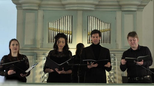 Vier Personen stehen mit Gesangsmappen vor einer Orgel und Singen.