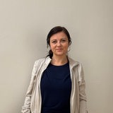 Eine Frau mit dunklen Haaren steht vor einer Wand und schaut in die Kamera