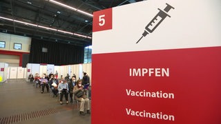 Ein Aufsteller mit der Aufschrift "Impfen" im Vordergrund. Im Hintergrund sind Personen, die warten, in der Messehalle zu sehen.