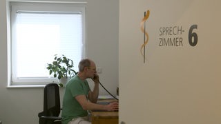 Hausarzt Holger Schelp sitzt am Bürotisch in seinem Sprechzimmer und telefoniert.