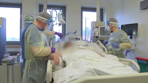 Zwei Ärzte stehen mit voller Schutzausrüstung um einen Patienten herum, der im Bett liegt und an Covid-19 erkrankt ist.
