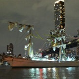 Ein Segelschiff mit abgeknickten Masten liegt vor einer Skyline an einer Brücke.