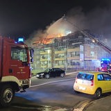 Ein brennendes Mehrfamilienhaus im Hintergrund, davor ein Feuerwehrwagen.