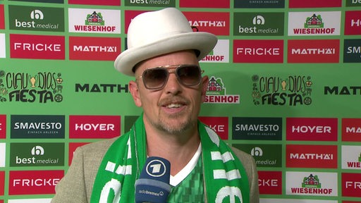 Rapper Jan Delay mit Hut und Sonnenbrille vor einer Werbewand bei Pizarros Abschiedsspiel im Interview.