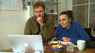 Ein Mann mit Bart und eine dunkelhaarige Frau schauen auf ein MacBook.