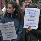 Mehrere Menschen stehen mit Schildern bei einer Demonstration vor einer Halle.