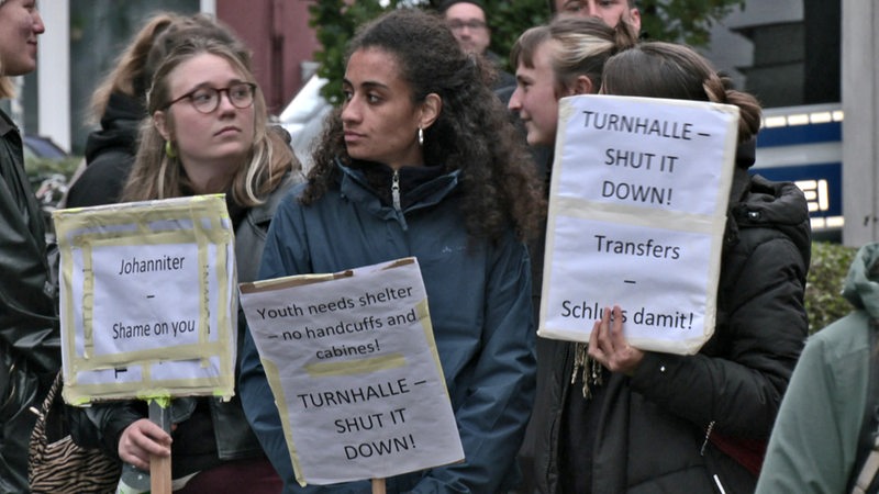 Mehrere Menschen stehen mit Schildern bei einer Demonstration vor einer Halle.