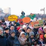 Mehrere Menschen lauschen einer Kundgebung auf einer Demonstration und halten Schilder hoch.