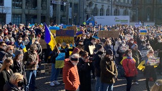 'Zahlreiche Menschen zum Teil mit Flaggen in den Farben der Ukraine blau und gelb stehen auf einem großen Platz, im Hintergrund ein historisches Gebäude