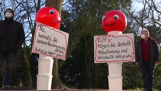Demonstrierende und zwei Figuren aus Plastikeimern und roten Luftballons. Sie tragen Schilder, auf denen etwas geschrieben steht.