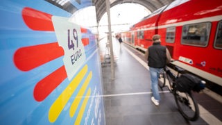 Ein Mann geht an einem Bahnhof mit seinem Fahrrad zwischen der Werbung für das Deutschlandticket und einer Regionalbahn vorbei.