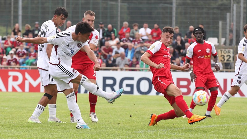 Ein Fußballer in einem weißen Trikot schießt, ein Gegenspieler in rot versucht, dies zu verhindern.