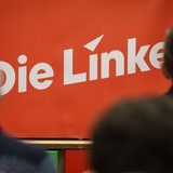 Menschen sitzen vor einem Logo mit der Aufschrift "Die Linke"