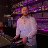 Ein junger Mann in Hemd steht hinter einer Bar und schaut in die Kamera