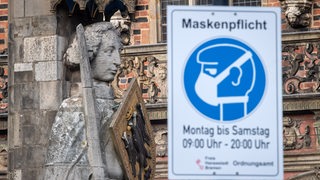 Ein Schild weist auf die Maskenpflicht hin. Im Hintergrund Bremer Roland und Rathaus.