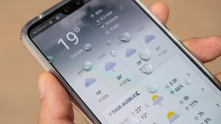 Ein Mann hält ein Smartphone in der Hand, das eine Wetter-App zeigt (gestellte Szene). 