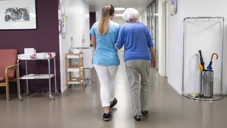 Eine Pflegerin führt eine ältere Dame durch einen Flur
