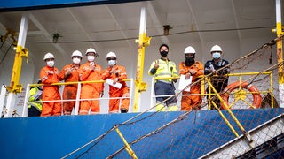 Mehrere Männer stehen mit Masken an der Rehling eines Schiffes.
