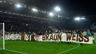 Spieler des FC St. Pauli halten ein Banner mit der Aufschrift "Hamburg ist braun-weiss" vor den Fanblock. 