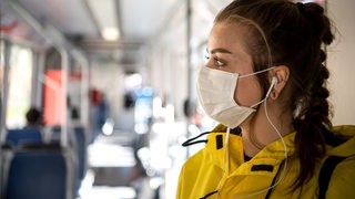 Eine junge Frau steht mit einer gelben Jacke bekleidet und einer Maske in einer Bremer Straßenbahn.
