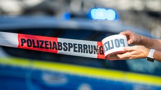 Ein Absperrband wird vor einem Polizeiwagen ausgerollt, an dem das Blaulicht leuchtet.