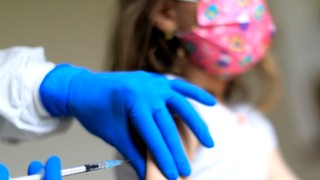 Symbolbild zeigt im Hintergrund ein Mädchen mit Maske, was gegen Covid geimpft wird