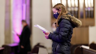 Eine junge Frau mit Maske und Winterkleidung singt vom Blatt in einer Kirche