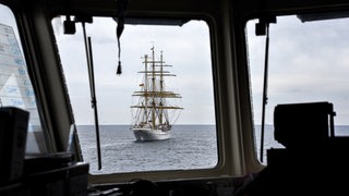 Durch das Fenster eines Schiffes ist ein Segelschiff auf dem Meer zu sehen.