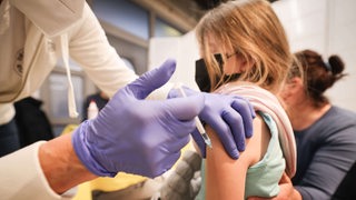 Zwei Hände mit Handschuhen geben einem Mädchen eine Impfung in den Arm.