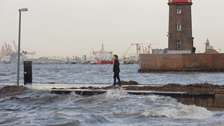 Eine Frau steht auf einer Mohle zwischen hohen Wellen vor dem Hafen.