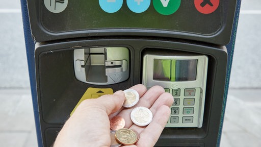 Eine Hand hält Euromünzen vor einem Parkscheinautomaten.