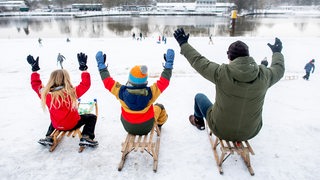 Drei Personen sitzen auf ihren Schlitten auf einem verschneiten Deich. Im Hintergrund sind ein Fluss und weitere Personen mit Schlitten zu erkennen.
