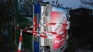 Ein Zigarattenautomat hängt zerstört an einem Metallzaun