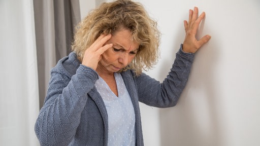 Eine ältere Frau mit Kopfschmerzen und Schwindel stützt sich an einer Wand ab
