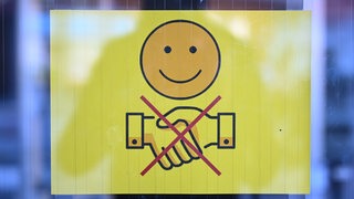 Ein Schild, das ein freundlich lächelndes Gesicht und zwei durchgestrichen Hände, die zum Gruß gereicht werden, zeigt, hängt an der Eingangstür eines Gymnasiums.