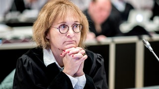 Eine Frau mit runder Brille und Anwaltsrobe sitzt mit verschränkten Händen im Gerichtssaal