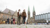 Ein junger Mann und eine junge Frau stehen Händchen haltend mit Maske vor Rathaus, Dom und Bürgerschaft Bremen.