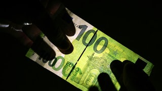Ein Hundert-Euro-Geldschein ist im Gegenlicht zu sehen. Zwei Hände halten ihn vor das Licht.