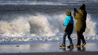 Eine Frau steht mit einem Kind am Strand vor den tosenden Wellen der Nordsee und macht ein Bild mit ihrem Handy. 