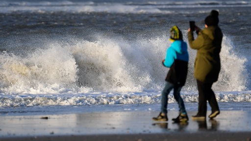 Eine Frau steht mit einem Kind am Strand vor den tosenden Wellen der Nordsee und macht ein Bild mit ihrem Handy. 
