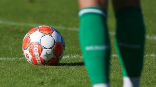 Ein Fußball liegt auf einem Rasen mit weißen Linien in Nahaufnahme, davor sieht man Beine eine Fußballspielers mit grünen Strümpfen.