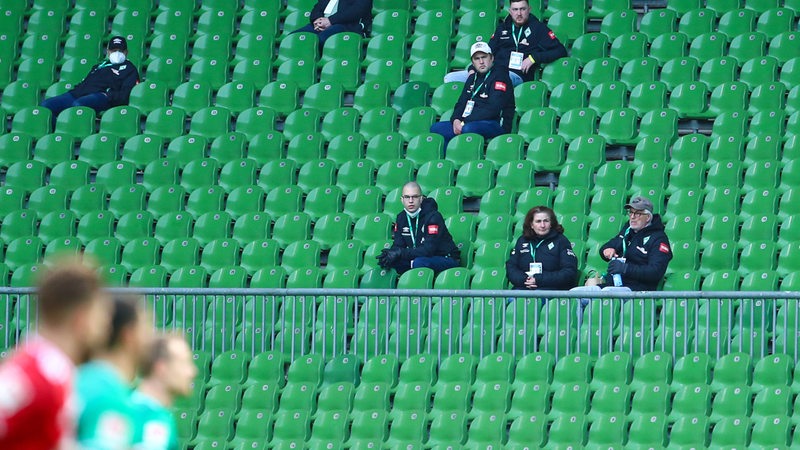 Auf einer Tribüne in einem Fußballstadion sitzen vereinzelt und jeweils allein wenige Menschen.
