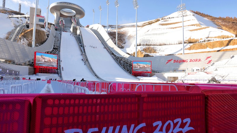 Blick von unten auf die olympische Skisprunganlage der Spiele in Peking im Sonnenschein mit Kunstschnee bedeckt.