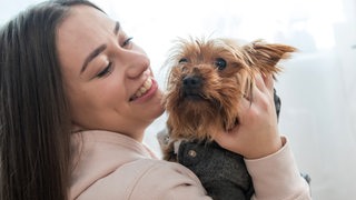 Eine junge Frau hält lachend ihren kleinen Hund auf dem Arm