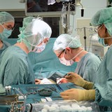 Ärzte und Op-Kräfte stehen in Kitteln und mit Masken an einem OP-Tisch, daneben ein Tisch mit Scheren und anderem OP-Besteck
