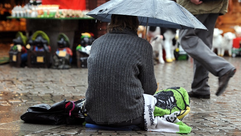 Ein Mann mit grauem Pullover sitzt im Regen auf dem gepflasterten Bode mit einem Schirm, im Hintergrund ein Stand mit Waren