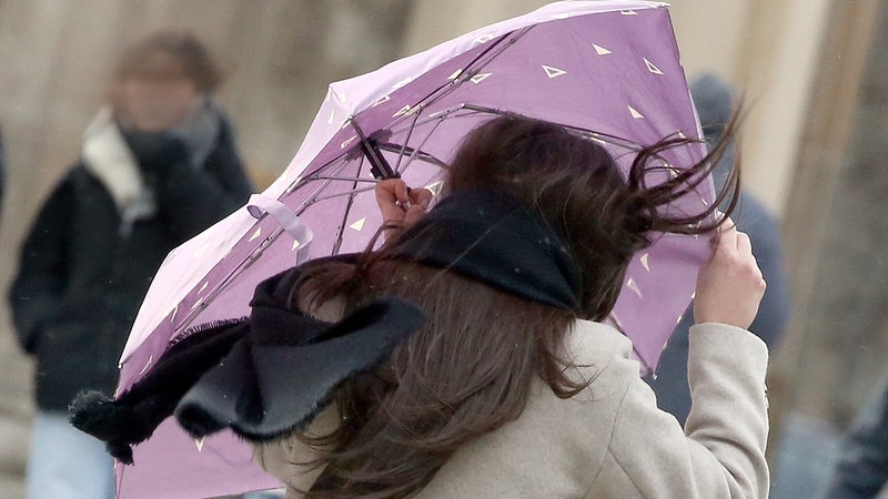 Eine Passantin mit wehenden Haaren sichert ihren Regenschirm, während sie sich bei Windstärken bis 100 km/h