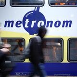 Ein Metronom steht im Bahnhof, Passagiere laufen vorbei