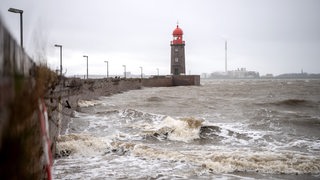 Wellen schlagen an eine Kaimauer. Im Hintergrund ist ein Leuchtturm zu sehen.